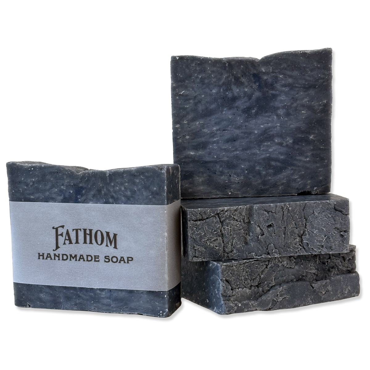 Four rough-cut bars of Fathom Handmade Soap in a dark charcoal gray color, with one bar wrapped in a silver paper band and two standing upright on a white background.