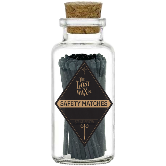 Glass jar with cork lid containing black safety matches labeled 'The Lost Wax Co.' on a white background.