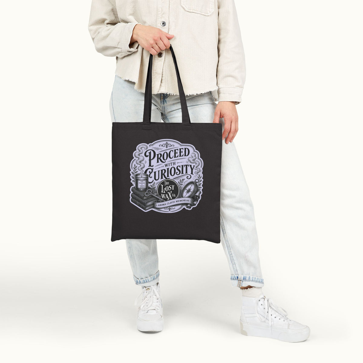 A person wearing a light beige jacket, light wash jeans, and white sneakers holds a black canvas tote bag with a vintage-style graphic that reads 'PROCEED WITH CURIOSTITY'.
