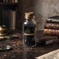 A vintage-style clear glass bottle filled with black safety matches, sealed with a cork stopper and labeled 'The Lost Wax Co. Safety Matches,' set on a rustic wooden table with old books and magnifying glasses in the background.