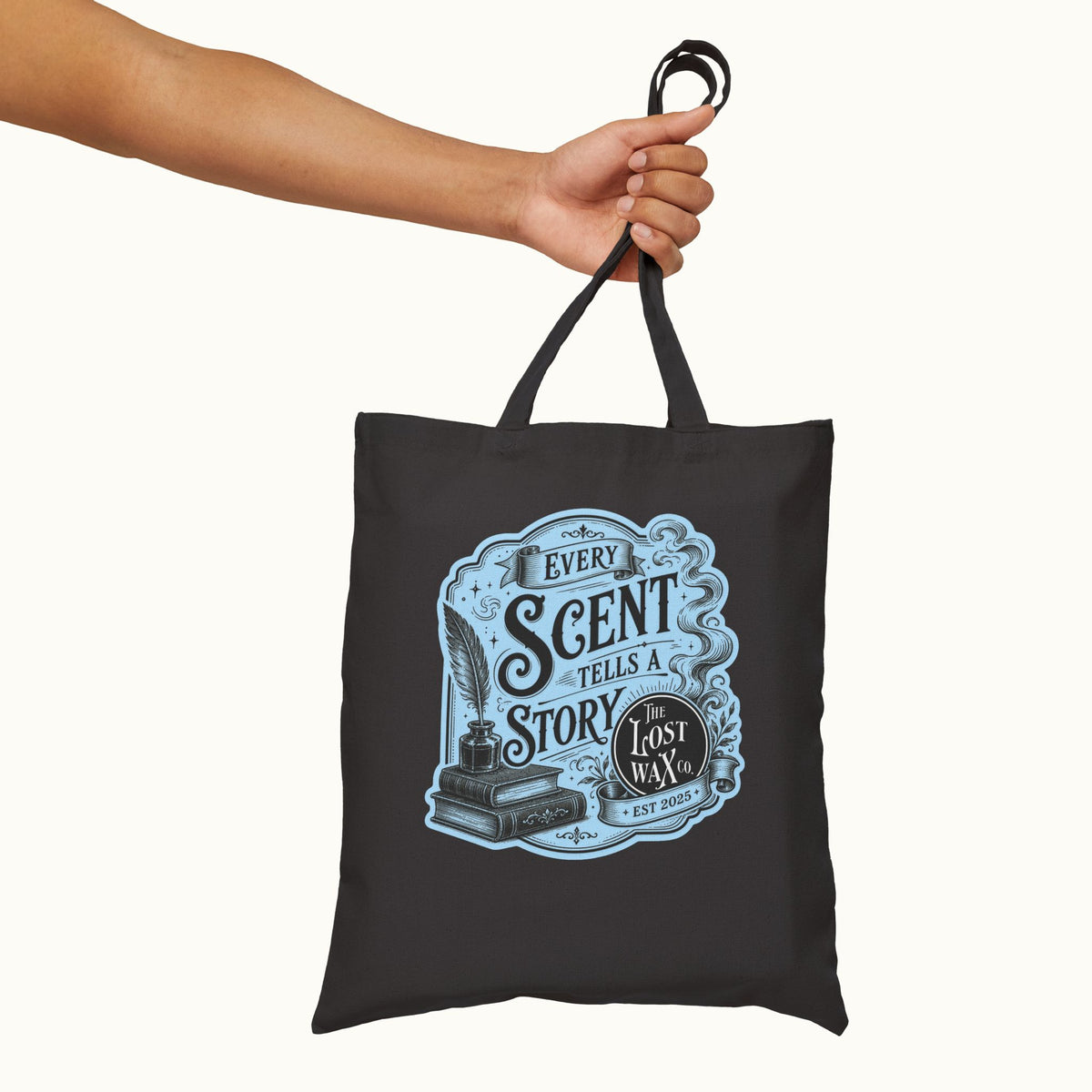 A black canvas tote bag with a light blue and white graphic print reading 'Every Scent Tells a Story' and 'The Lost Wax Est. 2025', held by a person's arm against a plain off-white background.