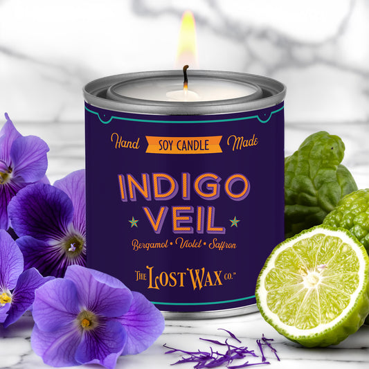 Candle in a can labeled 'Indigo Veil' by The Lost Wax Co., surrounded by purple flowers and a lime.