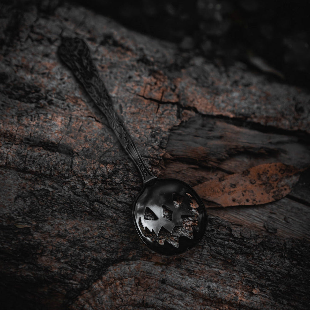A dark metallic spoon with a decorative handle and a jack-0-lantern-shaped cutouts in the bowl, resting on textured dark wood bark.