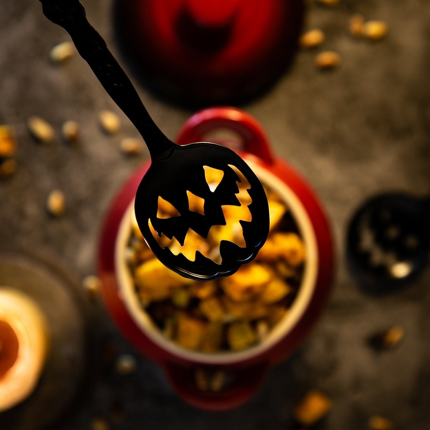 Black spoon with carved pumpkin design over a bowl of snacks on a dark background