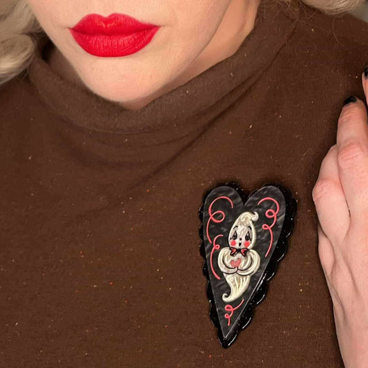 A close-up of a person wearing a brown turtleneck sweater and bright red lipstick, with a heart-shaped black brooch pinned to the chest. The brooch features a white and gray cartoon ghost with red details.