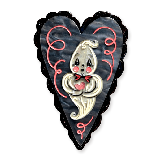 A heart-shaped brooch featuring a cute cartoon ghost with a red bow tie and pink heart, set against a dark marbled background with a black scalloped border.
