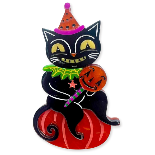 A Halloween-themed brooch featuring a black cat wearing an orange and pink pointed hat, holding a carved orange pumpkin, and sitting on a red pumpkin base. The cat has yellow eyes, a green collar with a spider detail, and holds a purple-striped candy cane.