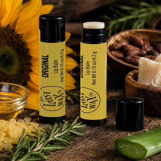 Two tubes of Lost Wax Co. Herbal Mint Lip Balm are displayed on a rustic wooden surface with natural ingredients including rosemary, aloe vera, cocoa beans, and a sunflower in the background.