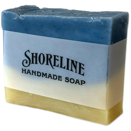 A rectangular handmade soap bar with a blue-to-cream-to-yellow gradient, featuring a white paper label that reads 'SHORELINE HANDMADE SOAP'.