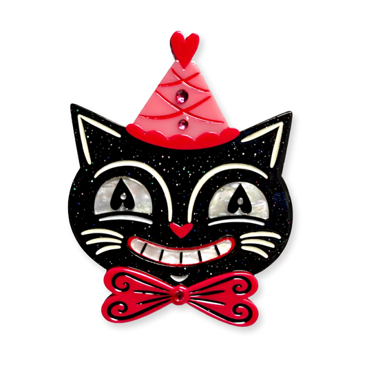A decorative black cat face with white and silver shell-like eyes, a red party hat, and a red bow tie on a plain white background.